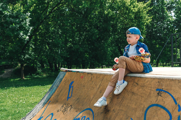 A small city boy and a skateboard. A young guy is standing in the park and holding a skateboard. City Style. City children. A child learns to ride a skateboard - Φωτογραφία, εικόνα