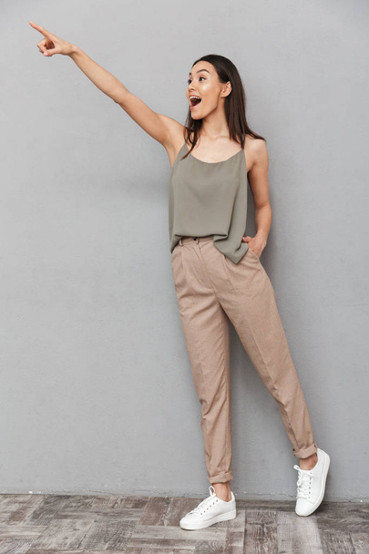 Full length portrait of an excited asian woman pointing finger away at copy space over gray background - Foto, immagini