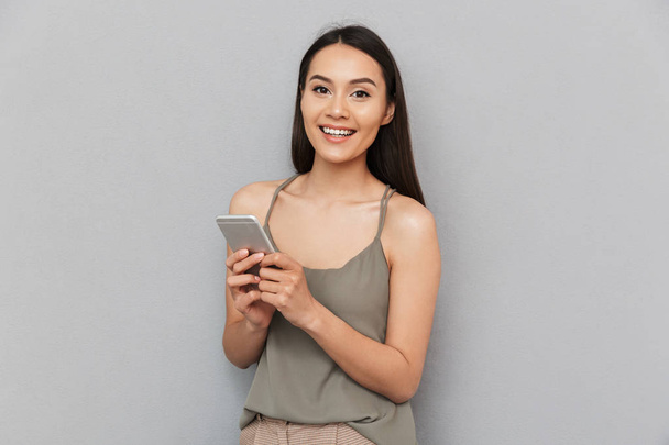 Portrait of a cheerful asian woman holding mobile phone and looking at camera isolated over gray background - Foto, Bild