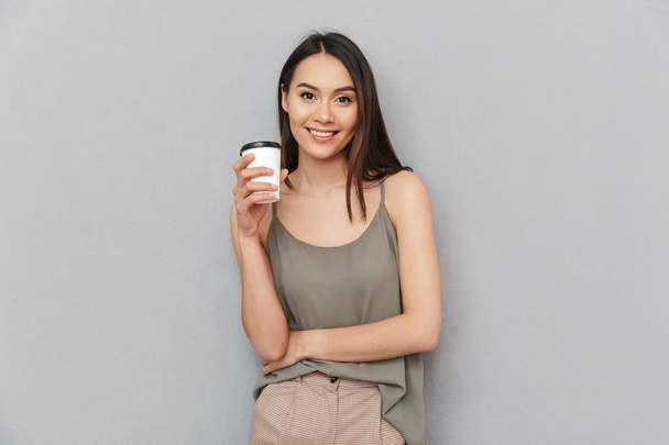 Portrait of a lovely asian woman holding take away coffee cup and looking at camera isolated over gray background - Photo, image