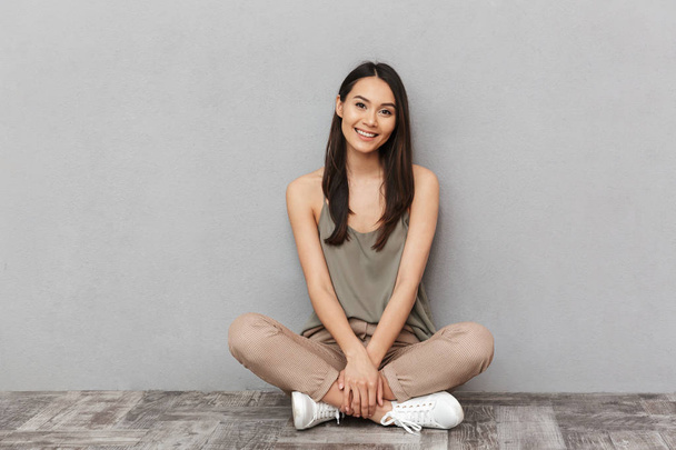 Portrait of a smiling asian woman sitting on a floor with legs crossed over gray background - Foto, afbeelding