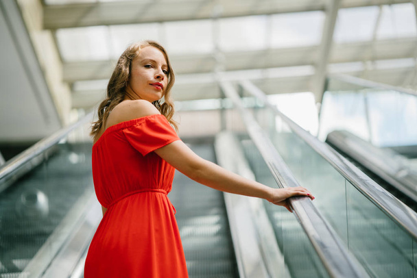 portrait of the beautiful young  woman  in  orange summer dress on escalator.  - Фото, изображение