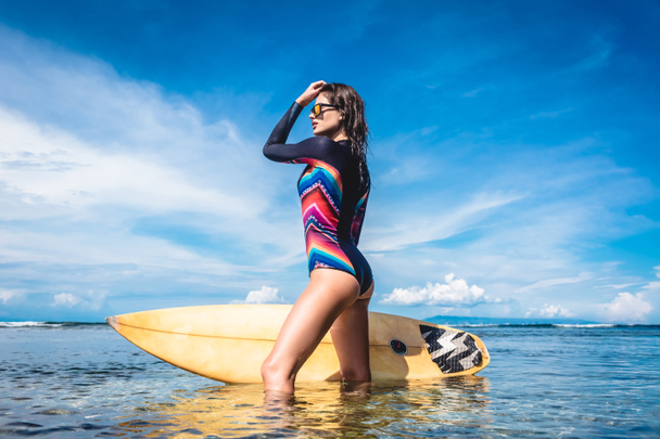 attractive young woman in wetsuit and sunglasses with surfboard posing in ocean at Nusa dua Beach, Bali, Indonesia - Photo, Image