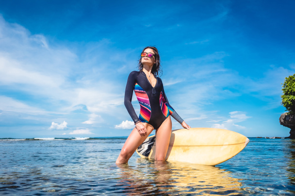 hermosa mujer joven en traje de neopreno y gafas de sol con tabla de surf posando en el océano en Nusa dua Beach, Bali, Indonesia
 - Foto, imagen