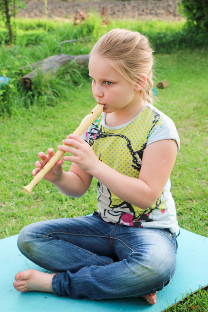 Portrait of beautiful little girl with blond hair in jeans playing flute in the garden sitting on the lawn, on a summer day - Photo, Image