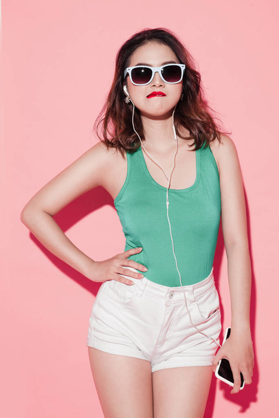 Jovem atraente mulher asiática em óculos de sol ouvindo música no fundo rosa
. - Foto, Imagem
