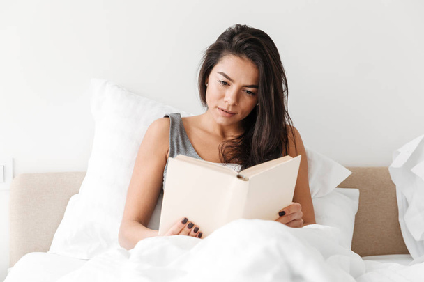 Portrait of relaxed adorable woman with long brown hair resting in comfortable bed after sleep and reading book - Photo, image