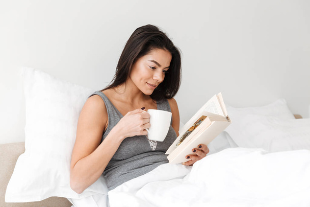 Portrait of pleased peaceful woman with long brown hair resting in comfortable bed after sleep with reading book and drinking coffee - Foto, Imagem
