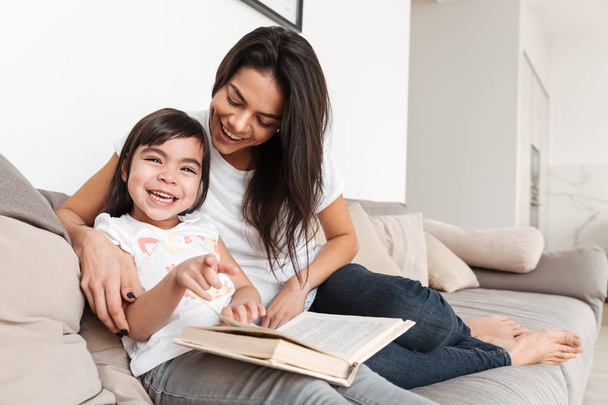 Portrait of joyful family mother and child spending time together sitting on sofa in living room and reading book - Фото, изображение