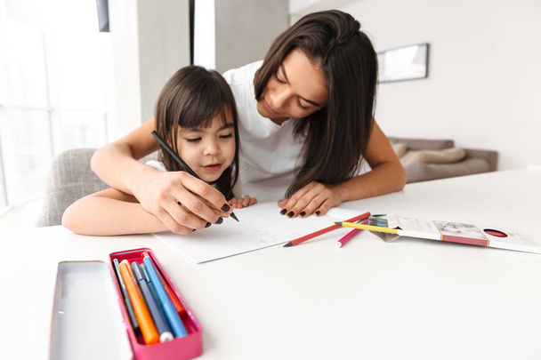 Image of beautiful people woman and child taking pleasure while sitting at table in flat and drawing picture with pencils together - Photo, image