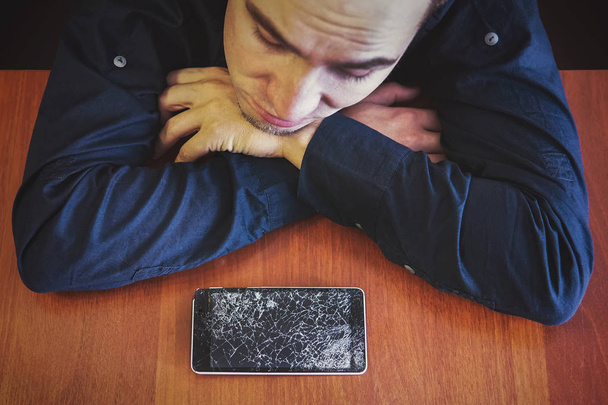Člověk se dívá na jeho rozbité telefon ležící na dřevěný stůl smutným pohledem. Guy byl rozrušený o prolomení jeho telefon. Rozbité telefon displej - Fotografie, Obrázek