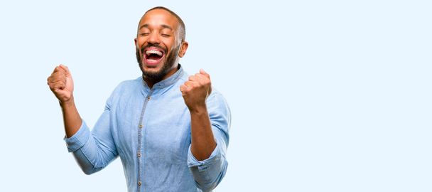 Hombre afroamericano con barba feliz y sorprendido animando expresando un gesto de sorpresa aislado sobre fondo azul
 - Foto, imagen
