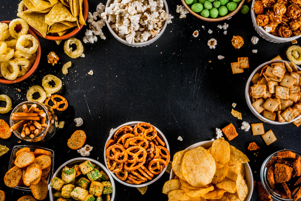 Variation different unhealthy snacks crackers, sweet salted popcorn, tortillas, nuts, straws, bretsels, back chalkboard copy space - Photo, Image