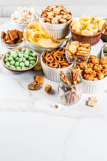 Variation verschiedene ungesunde Snacks Cracker, süß gesalzenes Popcorn, Tortillas, Nüsse, Strohhalme, Brezel, weißer Marmor Hintergrund Kopierraum - Foto, Bild