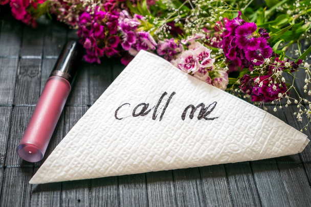 The inscription CALL ME on a white napkin left on the table in a cafe. Text call me on a background of pink flowers and lipstick. Dating, Love meeting  - Фото, изображение