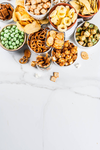 Variation verschiedene ungesunde Snacks Cracker, süß gesalzenes Popcorn, Tortillas, Nüsse, Strohhalme, Brezel, weißer Marmor Hintergrund Kopierraum - Foto, Bild