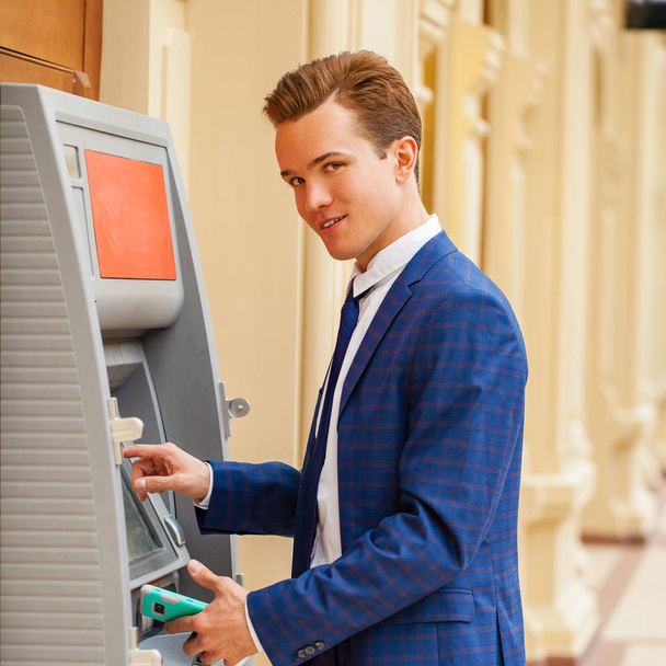 A young businessman in a blue suit stands against the background of an ATM in a shopping center - 写真・画像