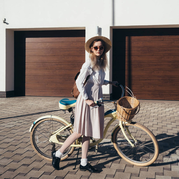 Pretty girl in hat and dress by cruiser bicycle in suburb. - Фото, зображення