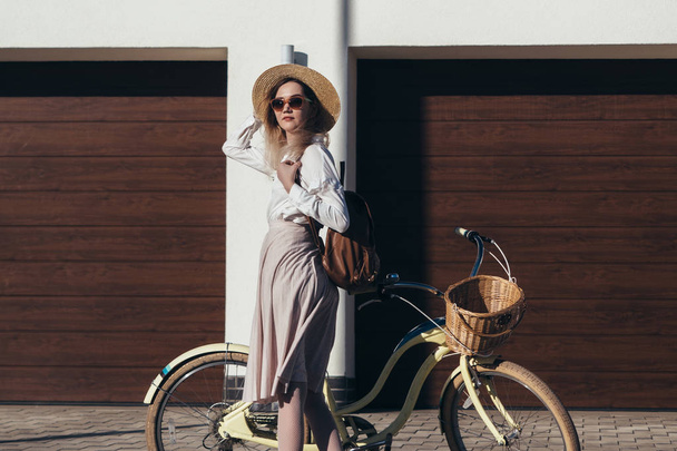 Pretty girl in hat and dress by cruiser bicycle in suburb. - Фото, изображение