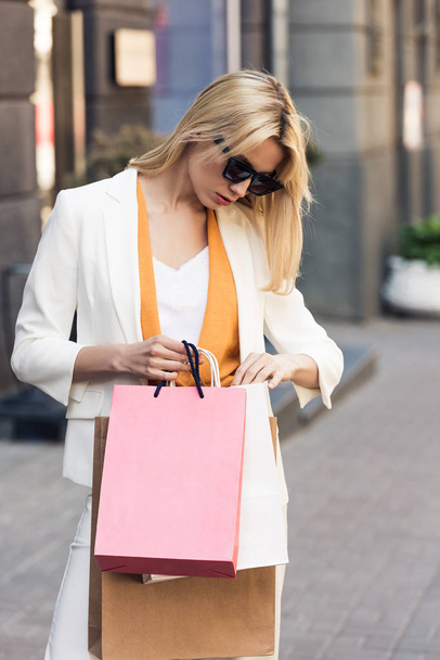 stylish young woman in sunglasses looking into shopping bags on street - Valokuva, kuva