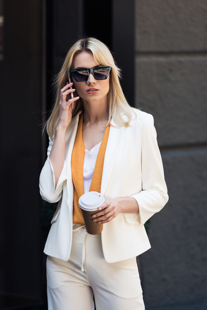 elegante mujer joven sosteniendo taza de café desechable y hablando por teléfono inteligente en la calle
 - Foto, imagen