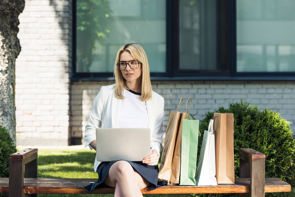 jeune femme blonde dans les lunettes à l'aide d'un ordinateur portable tout en étant assis sur le banc avec des sacs à provisions
 - Photo, image