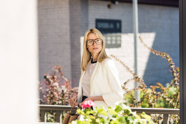 beautiful young blonde woman in eyeglasses holding shopping bags and looking away - Foto, Imagem
