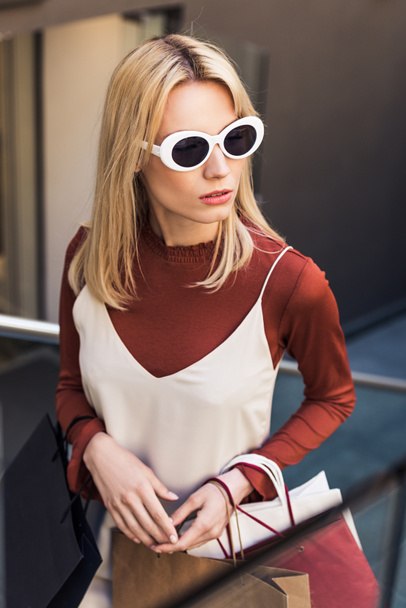 high angle view of beautiful young woman in sunglasses holding paper bags and looking away - Fotografie, Obrázek