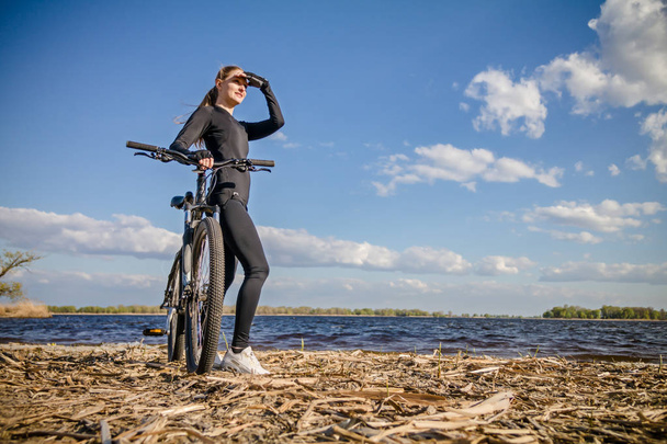 Νεαρή γυναίκα με ποδήλατο στέκεται στο riverside και αναζητούν μακριά - Φωτογραφία, εικόνα