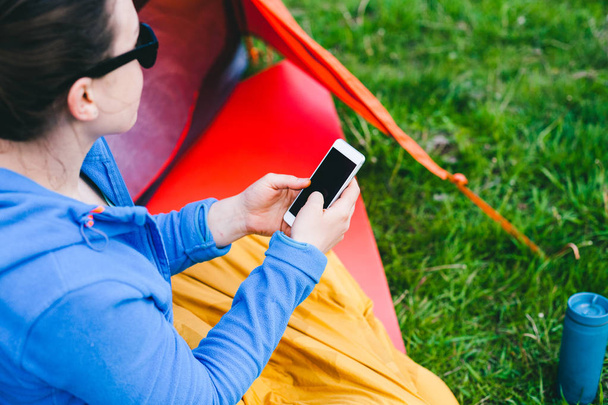 The girl is sitting near the tent. A woman is looking at the mobile phone and smiling. Tourist in a sleeping bag. Camping in nature. Mobile communication. - Фото, изображение