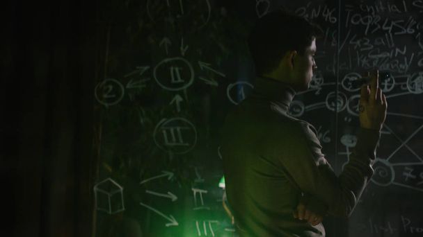 A man writes on a glass Board at night. Stock. An unrecognized genius alone writes down formulas on a blackboard - 写真・画像