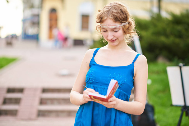 Όμορφο κορίτσι-καλλιτέχνης στον δρόμο, σε ένα μπλε φόρεμα γράφετε κάτι σε μια notebookl για οποιοδήποτε σκοπό - Φωτογραφία, εικόνα