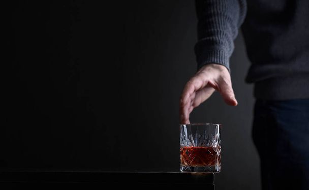 Man's hand reaches for a glass of alcohol. Conceptual image on the subject of alcoholism. Copy space for your text. - 写真・画像