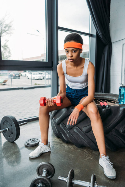mujer deportiva afroamericana en diadema y pulseras haciendo ejercicio con mancuerna mientras está sentada en el neumático de entrenamiento en el gimnasio
 - Foto, Imagen