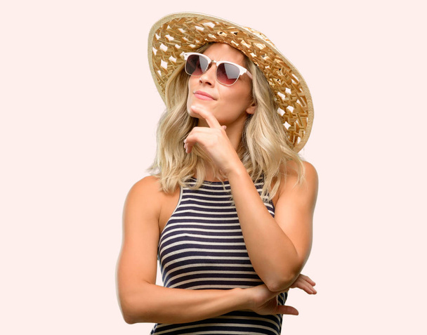 Jonge vrouw met behulp van zonnebril dragen zomer hoed denken en het opzoeken van uiten van de twijfel en verwondering - Foto, afbeelding
