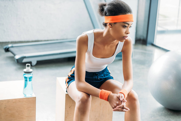 attractive african american sportswoman in headband and headbands resting on wooden box at gym - Foto, Imagen