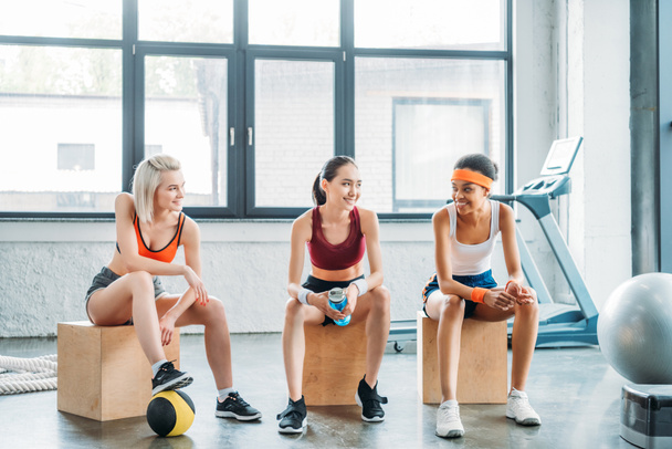 happy multiethnic female athletes talking to each other and sitting on wooden boxes at gym - Photo, Image