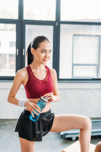 smiling asian sportswoman with bottle of water at gym - Фото, изображение
