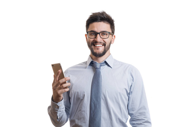 Young cheerful businessman in blue classic shirt with eyeglasses smiling and using his smartphone. Handsome bearded entrepreneur isolated on white background - Foto, imagen