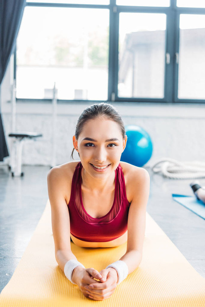 glimlachend Aziatische Sportvrouw in polsbandjes op fitness mat op sportschool - Foto, afbeelding