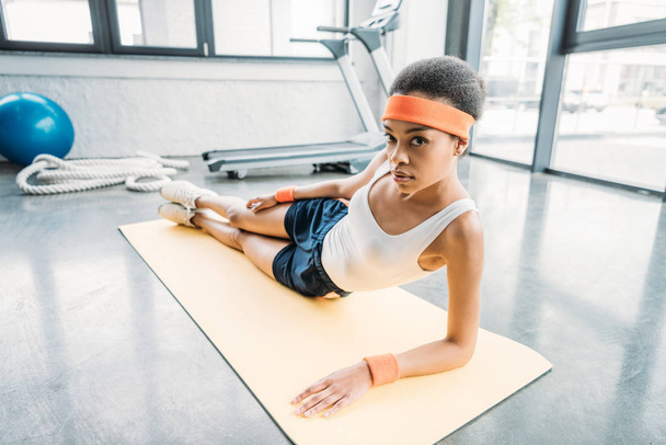 aantrekkelijke Afro-Amerikaanse atlete uit te oefenen op fitness mat en kijken naar camera op sportschool - Foto, afbeelding