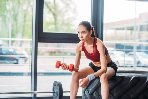 asian female athlete with dumbbell sitting on training tire at gym - Foto, imagen