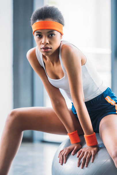 portrait of young african american sportswoman sitting on fitness ball at gym - Foto, Imagem