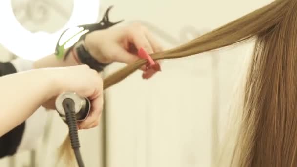 Hairstylist using hair tongs and comb for hairstyling in beauty salon. Close up hairdresser straightening long hair in hairdressing salon. - Filmati, video