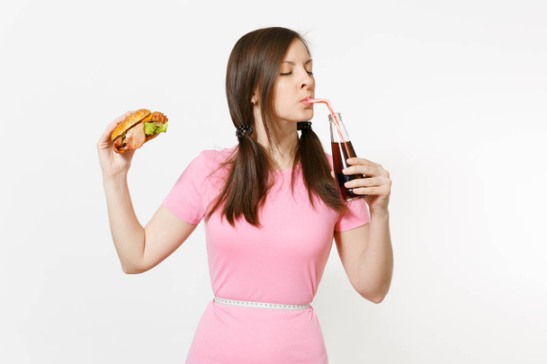 Fun young woman with tails, measuring tape at waist holding burger, cola in glass bottle isolated on white background. Proper nutrition or American classic fast food. Advertising area with copy space - Foto, Imagem