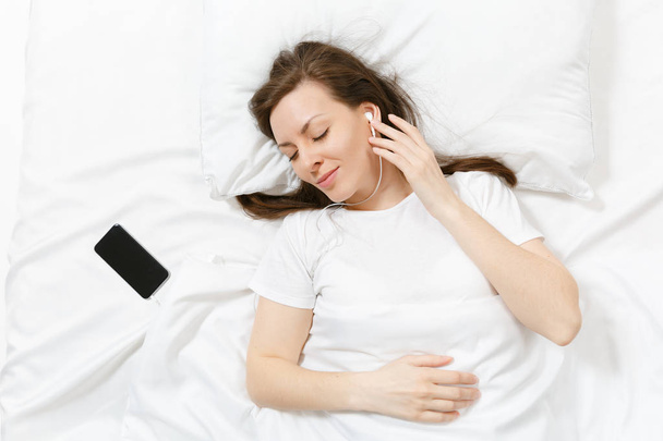 Top view of happy young woman lying in bed with white sheet, pillow, blanket, listen music from earphones in mobile phone. Calm beautiful female spending time in room. Rest, relax, good mood concept - Foto, imagen