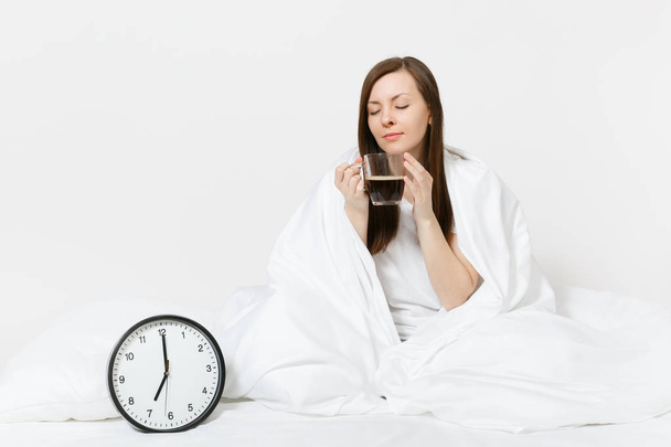 Happy woman sitting in bed with round clock, white sheet, pillow, wrapping in blanket on white background. Beauty female wake up early in morning, enjoys aroma of coffee. Rest relax good mood concept - Photo, Image