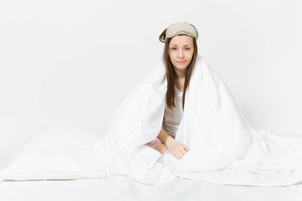Fun tired young woman sitting in bed with sleep mask, sheet, pillow, wrapping in blanket isolated on white background. Beauty female spending time in room. Rest, relax, good mood concept. Copy space - Photo, Image