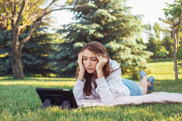 Portré, fiatal fehér kaukázusi nő fű néz digitális tábla parkban fekvő. Diák lány aggódik érzés hangsúlyozta. Erős érzelem, vicces arc kifejezése - Fotó, kép
