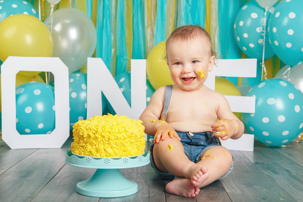 Portrait of cute adorable Caucasian baby boy in jeans pants celebrating his first birthday. Cake smash concept. Child kid sitting on floor in studio eating tasty yellow dessert  - Photo, image
